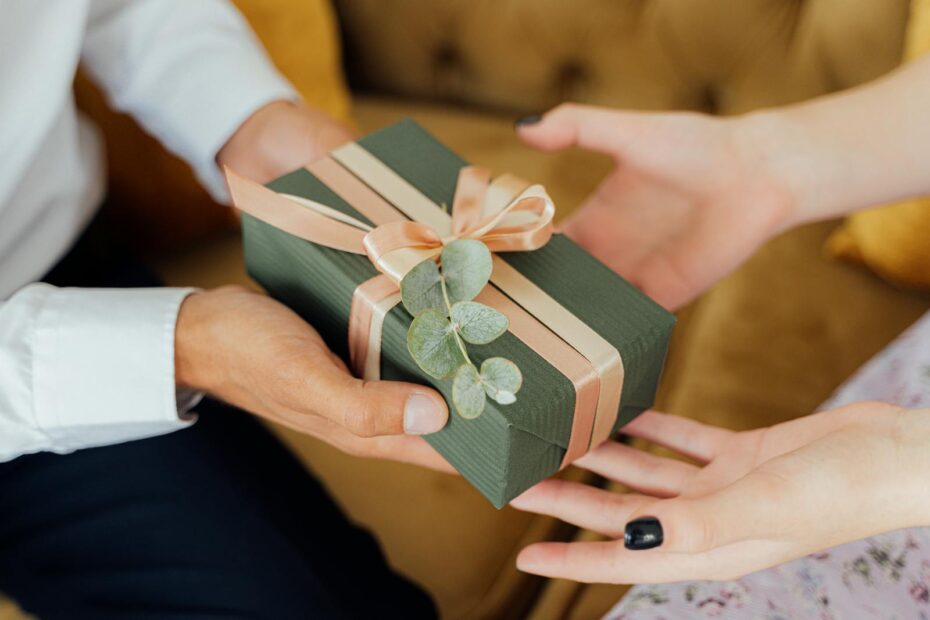 Two hands exchanging an elegantly wrapped green gift box with ribbons and foliage.