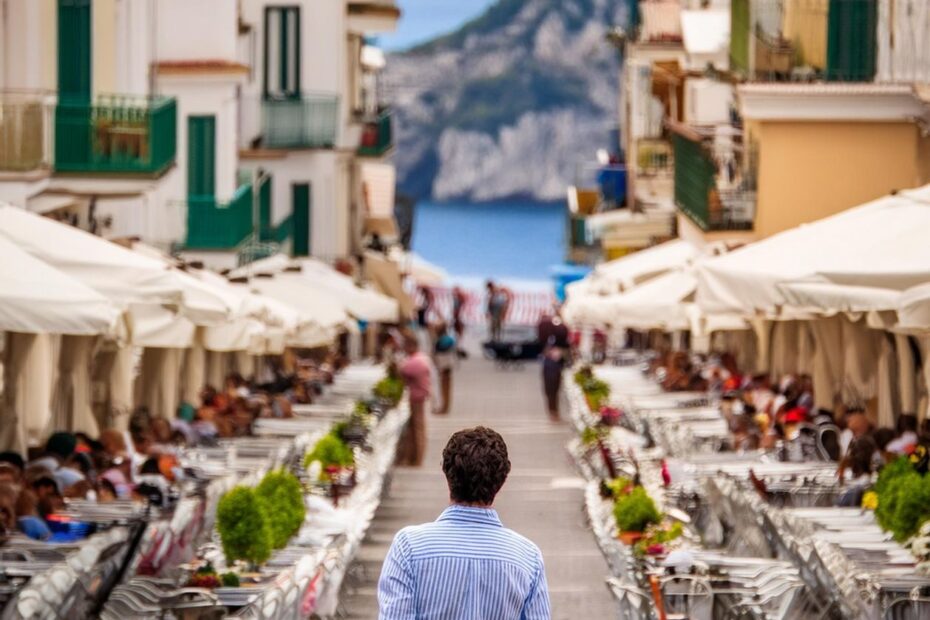 A man walks down a vibrant seaside street lined with cafés, with a scenic ocean view.